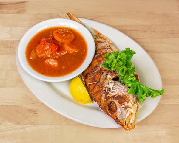 Whole fried red snapper served with a side of hearty vegetable stew and a lemon wedge, garnished with fresh lettuce on a white plate — a classic Caribbean seafood dish.