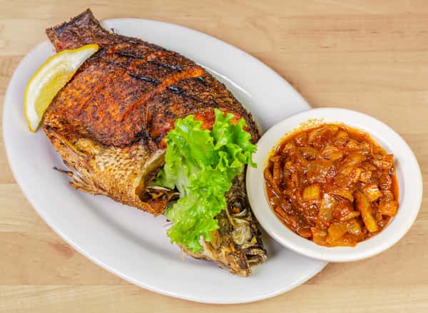Grilled whole tilapia seasoned with bold spices, garnished with fresh lettuce and lemon wedge, served alongside a bowl of savory tomato and onion sauce—popular in West African cuisine.