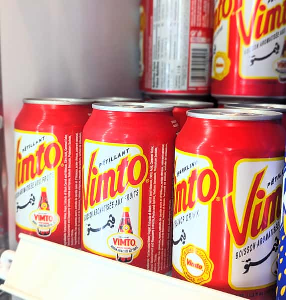 Assorted fruit-flavored beverages including red cans of sparkling Vimto and blue cans of Vani Float in mango and peach flavors, stocked in a refrigerated display.