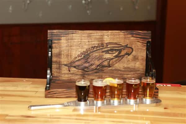 Flight of 5 beers in front of a wooden shelf with an eagle painted on it.