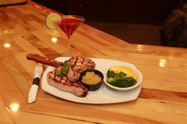 Steak with a side of broccoli on a white plate with a red martini with a lime slice on a wooden table.