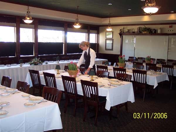 Inside banquet room set for event with potted flowers on the table. Female staff member is setting the table.