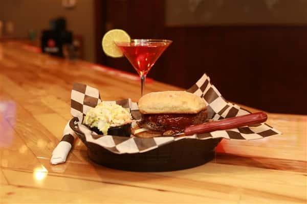 Sandwich with beef and covered in BBQ sauce and a side pickle spear. Red martini with lime slice on the side.