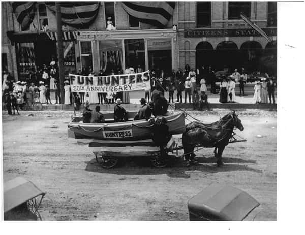 Black and white image of two horses pulling a boat on wheels in a parade.
