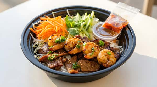 Takeout bowl with grilled shrimp and sliced beef over rice, with shredded carrots, greens, and sauce on the side.