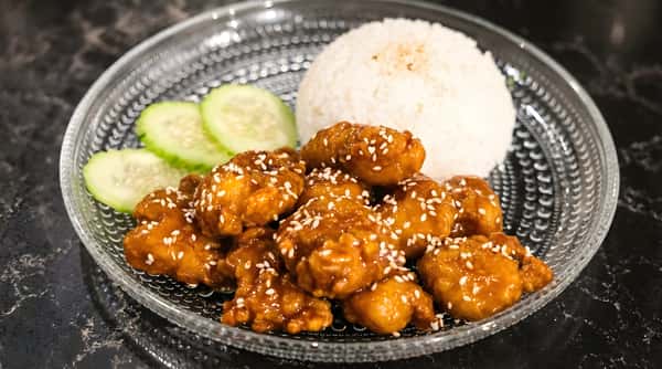 Orange chicken served with white rice and cucumber slices on a black plate.