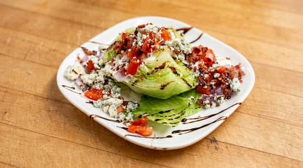 Wedge salad with iceberg lettuce, blue cheese crumbles, diced tomatoes, bacon bits, and balsamic glaze.