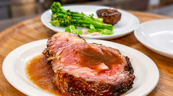 Slice of prime rib with gravy on a white plate, with green vegetables blurred in the background.