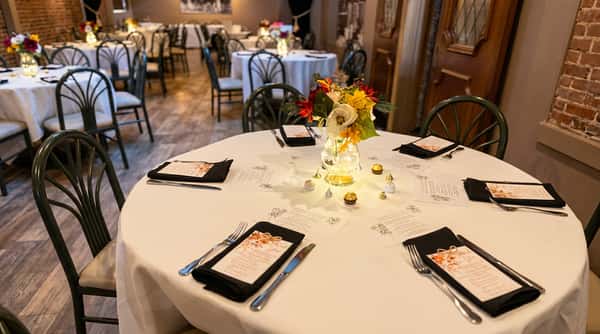 A white-covered dining table set with place settings, menus, a floral centerpiece, and chocolates in a dimly lit restaurant.