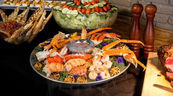 Seafood platter with lobster tails, crab legs, and shrimp on ice with dip, beside grilled bread, salad, and roast beef.