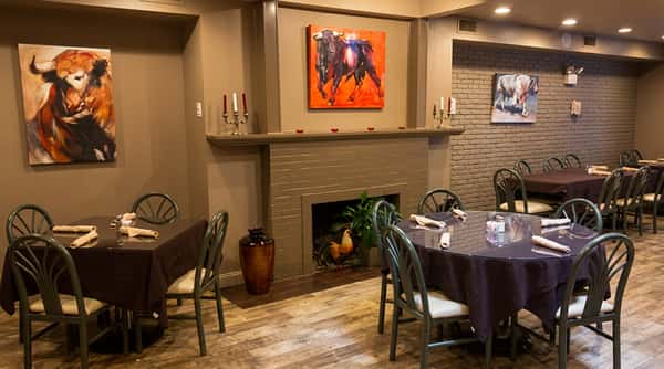 Restaurant dining room with bull paintings, a brick fireplace, and tables with dark tablecloths.