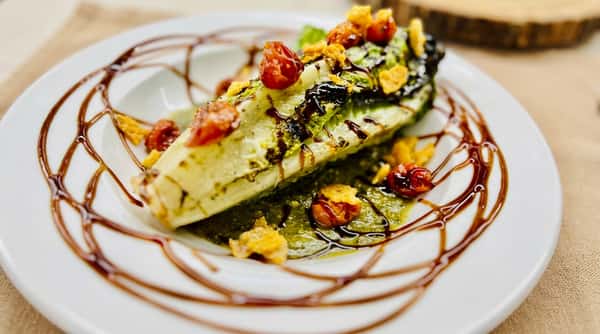 Grilled romaine heart with cherry tomatoes, croutons, green pesto, and balsamic glaze on a white plate.