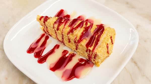 Layered dessert wedge topped with finely crushed crumbs, drizzled with red fruit sauce and white glaze.