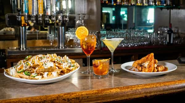 Nachos with jalapeños and white sauce, two cocktails, and chicken and waffles on a bar counter.