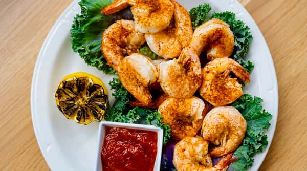 Seasoned shrimp on kale, with a grilled lemon half and a white ramekin of red sauce.