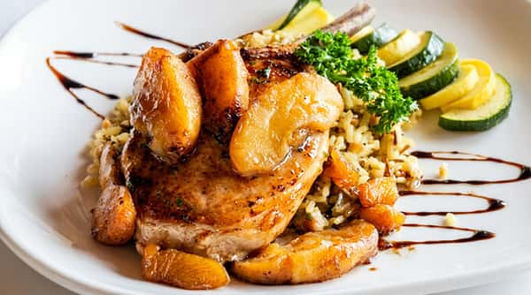 Pan-fried pork chop with glazed pears, wild rice, and sliced zucchini, drizzled with balsamic reduction.