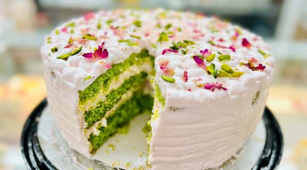 Pistachio cake with a slice removed, showing green cake layers with white frosting, topped with pink frosting, pistachios, and rose petals.
