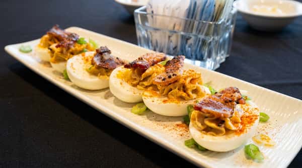 Deviled eggs on a white platter, topped with crispy bacon, paprika, and chopped green onions.