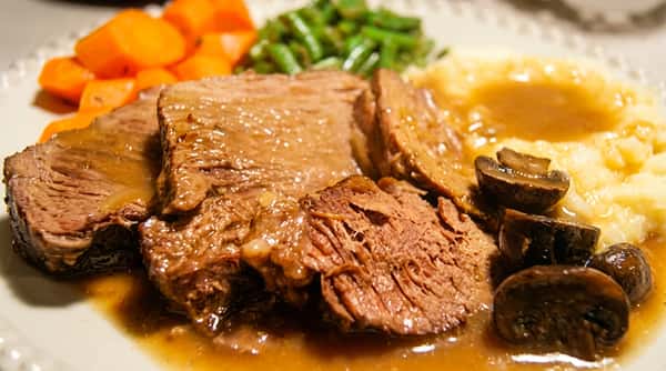 Pot roast with gravy, mashed potatoes, sliced carrots, green beans, and mushrooms.