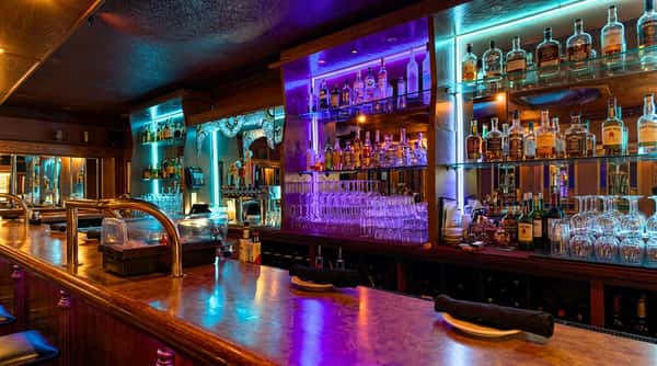 A bar lit with neon blue and purple lights, featuring rows of liquor bottles and wine glasses.