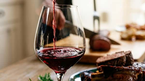 Red wine being poured into a clear glass, with a blurred background showing sliced meat and a carving knife.
