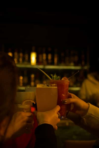 Friends clinking colorful cocktails and beer glasses in a dimly lit bar with a glowing bottle-lined shelf in the background.