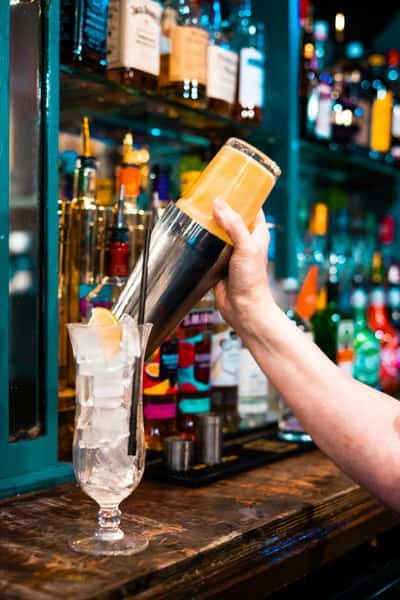 Bartender pouring a vibrant orange cocktail from a shaker into a tall ice-filled hurricane glass at a colorful bar, surrounded by an array of liquor bottles — ideal for promoting craft cocktails and nightlife ambiance.