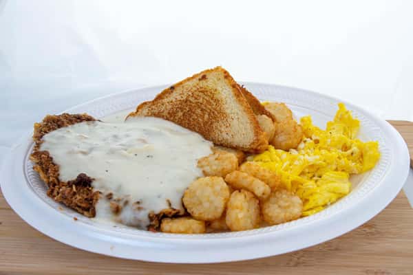 Country-fried steak with white gravy, scrambled eggs, tater tots, and toast on a white plastic plate.