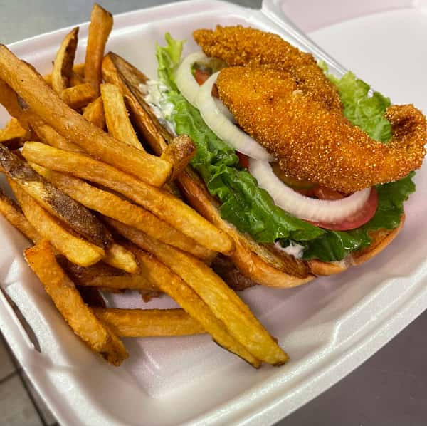 Fried fish sandwich topped with lettuce, tomato, and onion, served with golden fries in a foam takeout container.