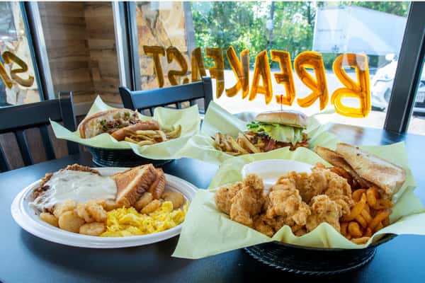 A table with three meals: a plate with scrambled eggs, tater tots, toast, and sausage gravy, a basket with fries and a sandwich, and a basket with fried chicken, fries, and toast, all in front of a window with "BREAKFAST" written on it.