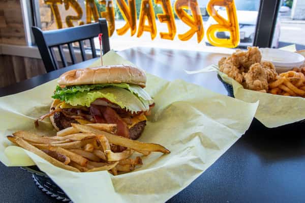 Bacon cheeseburger with lettuce, tomato, and onion served with fries in a basket, with chicken tenders and curly fries in the background.