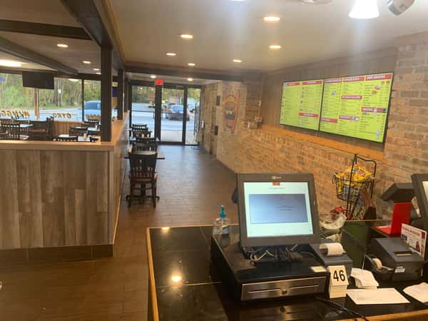 Front counter view of a casual restaurant with a touchscreen register, menu boards on a brick wall, and dining area with large windows.
