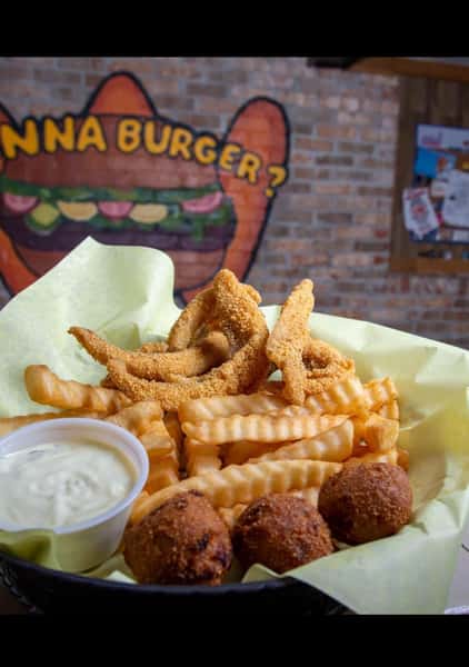 Basket of fried catfish strips, crinkle-cut fries, hush puppies, and tartar sauce served on yellow paper with a mural background.