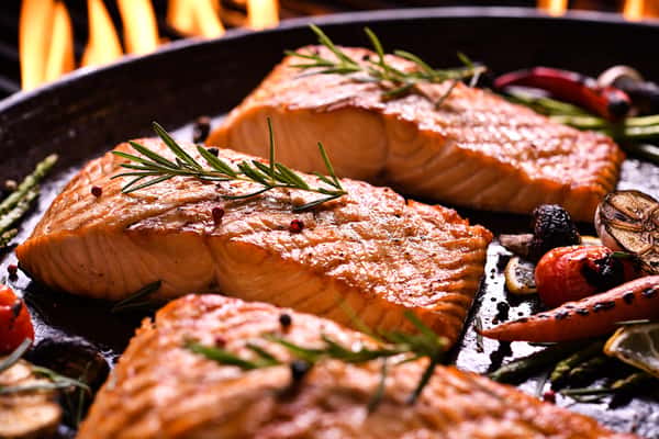 Grilled salmon fillets garnished with rosemary and served with roasted vegetables, with flames in the background.