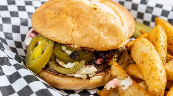Burger with picked jalapeños and white cheese on a checkered paper-lined basket with potato wedges.