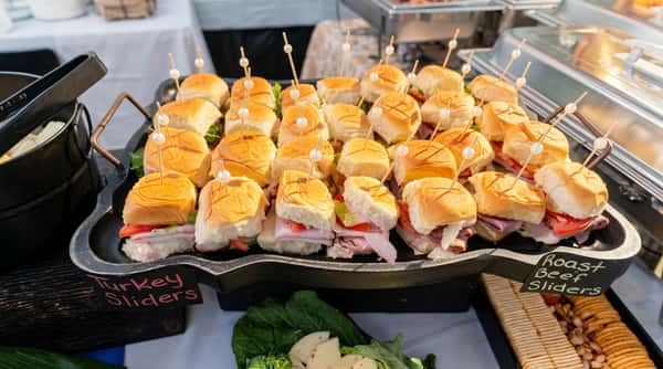 Platter of turkey and roast beef sliders with pearl-tipped toothpicks, alongside crackers, cheese, and broccoli.