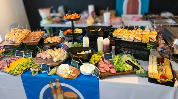 A buffet table laden with various appetizers: ham, turkey, and roast beef sliders, cheese boards, fruit, crackers, salami, and deviled eggs. Several black display boards organize and label the food items.