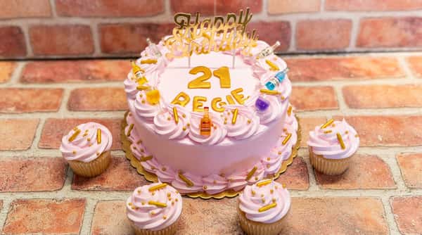 Pink birthday cake with gold "Happy Birthday," "21 REGIE" letters, and miniature liquor bottle/glass decorations. Four matching cupcakes are arranged around it.