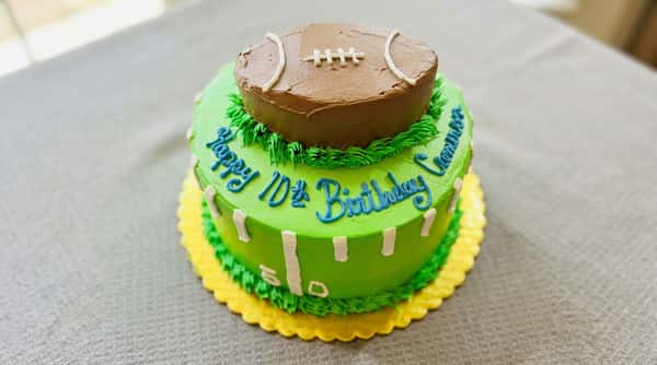 Two-tiered green and yellow football-themed cake with a brown football on top and "Happy 10th Birthday" in blue icing.