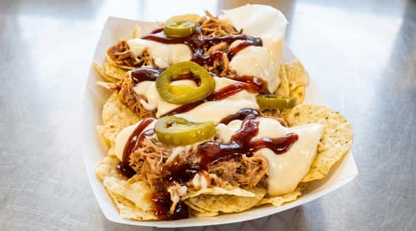 Pulled pork nachos with cheese sauce, BBQ drizzle, and pickled jalapeño slices in a white paper tray.
