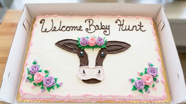 White sheet cake decorated with a brown and white cow face, pink and purple roses, and green leaves. Black icing spells "Welcome Baby Hunt."