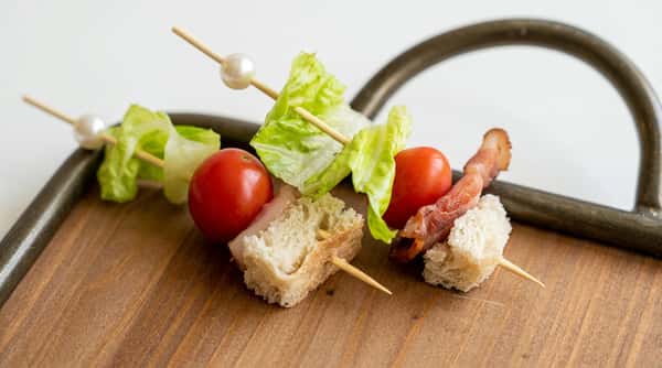 Appetizer skewers with cherry tomatoes, lettuce, slivers of prosciutto, and bread cubes on a wooden board.