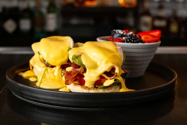 eggs Benedict on a black plate with a cup of fruit behind it