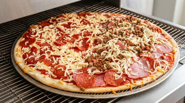 Uncooked pizza with pepperoni, ham, crumbled sausage, and shredded mozzarella cheese on a metal tray in an oven.