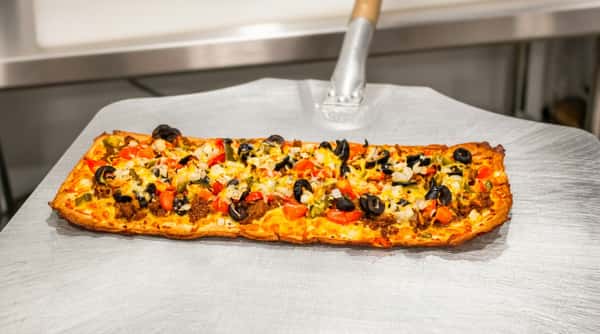 Rectangular flatbread pizza topped with ground meat, melted cheese, black olives, diced tomatoes, and onions on a metal peel.