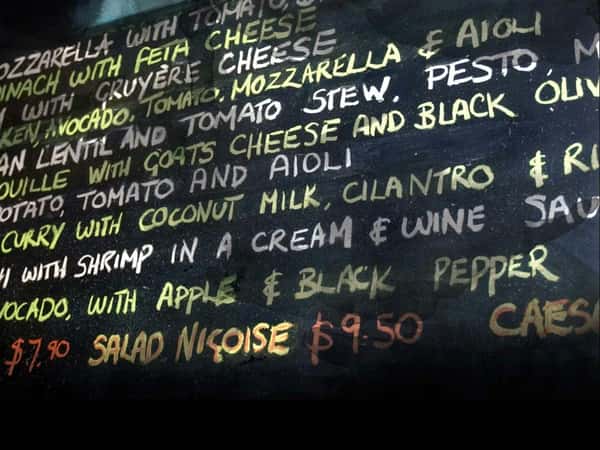 Close-up of a handwritten restaurant chalkboard menu featuring vibrant text listing dishes with ingredients like mozzarella, goat cheese, pesto, avocado, coconut milk, and black pepper, along with prices such as "$7.90" and "$9.50" for Salad Niçoise.