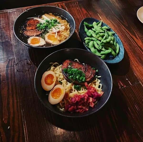 Top-down view of a Japanese meal featuring two bowls of ramen—one with broth, soft-boiled eggs, chashu pork, mushrooms, and green onions, the other a dry-style ramen with bacon bits, eggs, and charred pork—served alongside a plate of steamed edamame on a rustic wooden table.