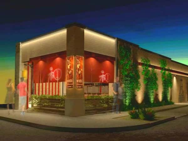 Exterior view of a modern Asian restaurant at dusk, featuring red signage with Japanese characters, warm ambient lighting, and green vertical plant accents on a brick facade, with blurred figures walking by.