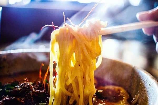 Steaming hot ramen noodles lifted by chopsticks from a rich, flavorful broth, with hints of sliced meat, scallions, and chili threads in a cozy, dimly lit setting.