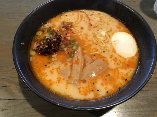 Bowl of spicy miso ramen in a rich, orange-toned broth, topped with a soft-boiled egg, bamboo shoots, ground meat, green onions, and chili oil, served in a black bowl on a wooden table.